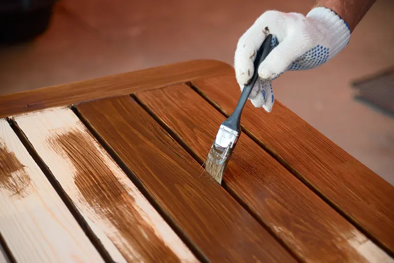 Mano con guante aplicando barniz oscuro sobre una mesa de madera, parte del servicio de terminaciones para muebles de madera de Ferrenova.