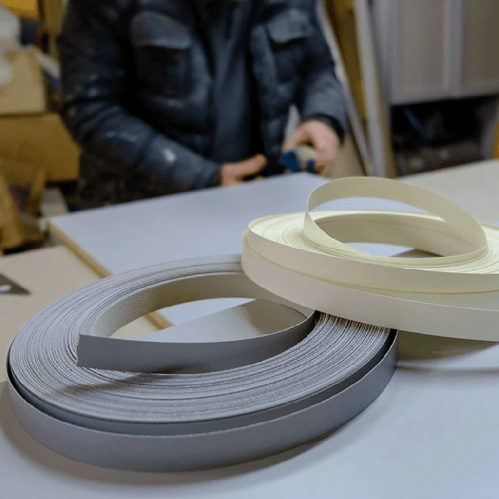 Rollos de cinta de PVC para enchapado de cantos de color gris y blanco sobre tablero de melamina, mostrando los materiales utilizados en el Servicio de enchapado de cantos profesional de muebles.