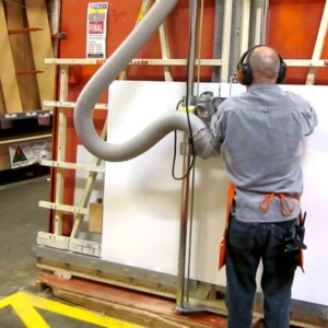 Trabajador con equipo de protección utilizando una sierra vertical industrial para realizar Cortes de melamina precisos en un gran tablero de color blanco.