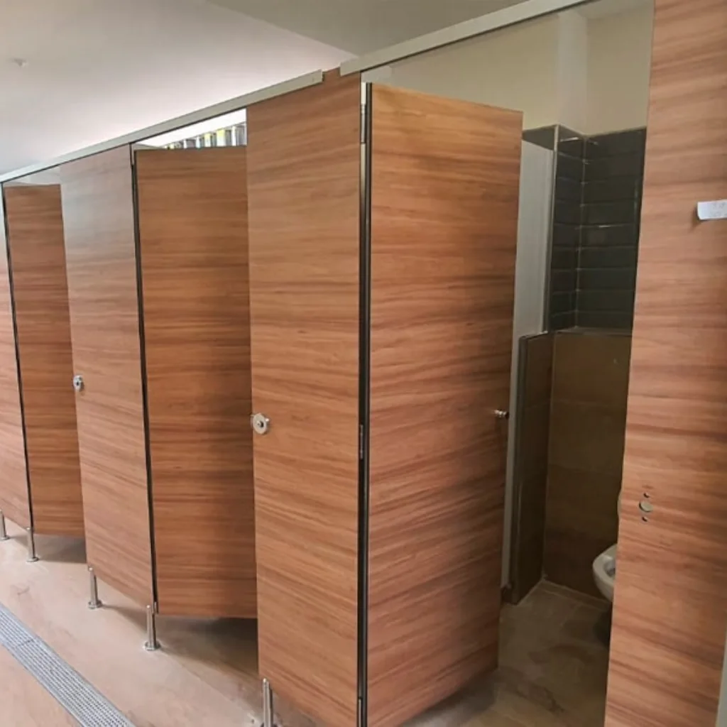 Filas de cabinas de baño con puertas y tabiques de HPL aspecto madera, elevadas del suelo y con herrajes de acero inoxidable en un sanitario público.
